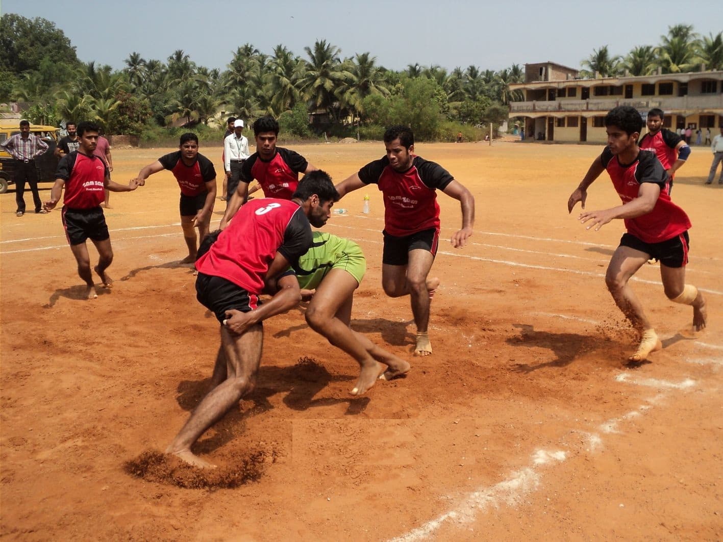 Kabaddi players contest a raid on a chalk-marked mud court, a defender diving in as the raider turns for the midline.